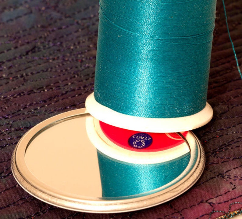 May include: A spool of teal thread with a white plastic bobbin and a red plastic base with the text "COV12" printed on it. The spool is sitting on a silver-colored, round, mirrored surface.