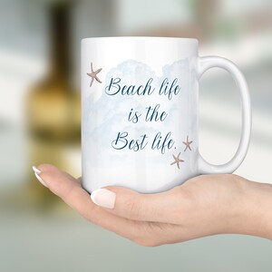 Peut inclure: Une tasse en c&eacute;ramique blanche avec un motif aquarelle bleu de nuages et le texte "Beach life is the best life." avec deux petites &eacute;toiles de mer brunes.