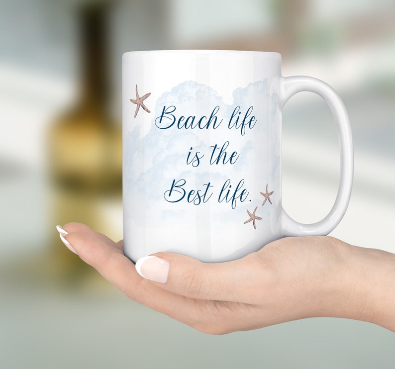 Peut inclure: Une tasse en c&eacute;ramique blanche avec un motif aquarelle bleu de nuages et le texte "Beach life is the best life." avec deux petites &eacute;toiles de mer brunes.