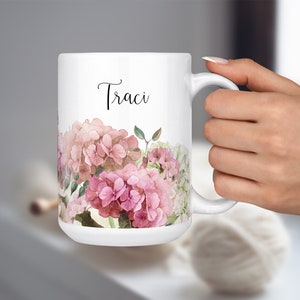 Op de afbeelding: Witte keramische mok met een bloemenontwerp met roze hortensia's en groene bladeren. De mok heeft de naam "Traci" in zwarte, cursieve letters.