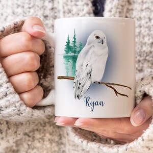 Peut inclure: Mug en céramique blanche avec une illustration à l'aquarelle d'un harfang des neiges perché sur une branche. Le design comprend une scène de forêt et le nom "Ryan".