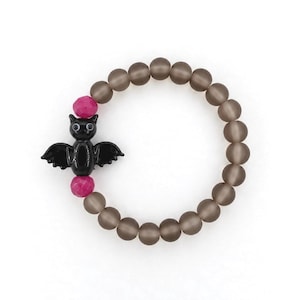 May include: A circular stretch bracelet featuring matte translucent grey beads, two faceted hot pink beads, and a central glossy black bat charm with white eyes. The bat is positioned between the two pink beads on the beaded strand.