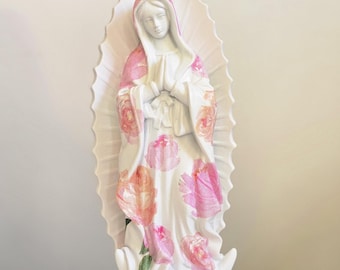 May include: A white statue of the Virgin Mary with a pink and white floral design on her robe. The statue has a halo and is standing on a white base. Religious home decor.