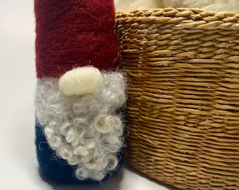 Könnte beinhalten: Eine handgefertigte Zwergpuppe mit roter Mütze, weißem Bart und blauem Körper. Der Zwerg steht neben einem geflochtenen Korb mit weißer Wolle. Der Zwerg ist eine festliche Dekoration.
