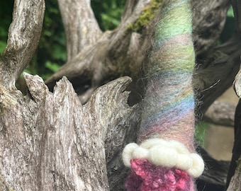 Könnte beinhalten: Eine handgefertigte Zwergenfigur mit einem konischen Hut in Grün-, Blau- und Rosatönen. Der Körper des Zwergs ist ein leuchtendes Rosa und er hat einen weißen Bart. Der Zwerg ist aus Wolle gefertigt und vor einem natürlichen Holzgrund platziert.