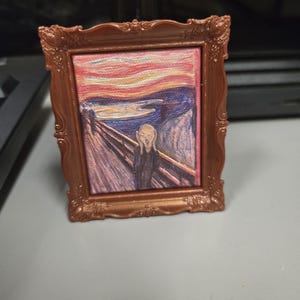 May include: A miniature framed artwork of Edvard Munch's "The Scream." The painting shows a figure with an open mouth against a backdrop of a fiery sky and a bridge. The frame is a decorative bronze colour.