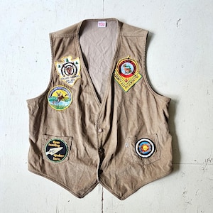 Może przedstawiać: Beżowa kamizelka sztruksowa z wieloma naszywkami. Naszywki zawierają tekst i obrazy, w tym jelenia, tarczę i słowa "Bow hunting spoken here". Kamizelka ma dekolt w serek i zapięcie na guziki.