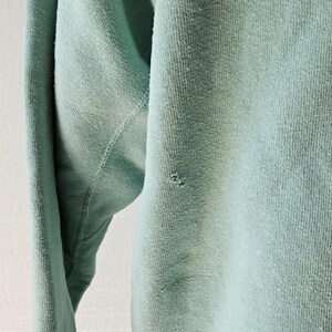 Op de afbeelding: Close-up van een lichtblauwe sweatshirtmouw. De stof vertoont tekenen van slijtage, met een klein gaatje bij de elleboog. De textuur van het gebreide materiaal is zichtbaar, wat wijst op een vintage of versleten kledingstuk.