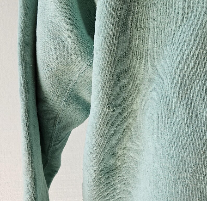 Op de afbeelding: Close-up van een lichtblauwe sweatshirtmouw. De stof vertoont tekenen van slijtage, met een klein gaatje bij de elleboog. De textuur van het gebreide materiaal is zichtbaar, wat wijst op een vintage of versleten kledingstuk.