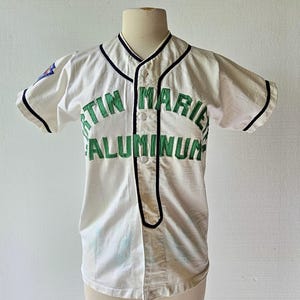 May include: White baseball jersey with black trim and green lettering that reads "TIN MARIE ALUMINUM". The jersey has short sleeves and a button-up front. A small patch is visible on the left sleeve.
