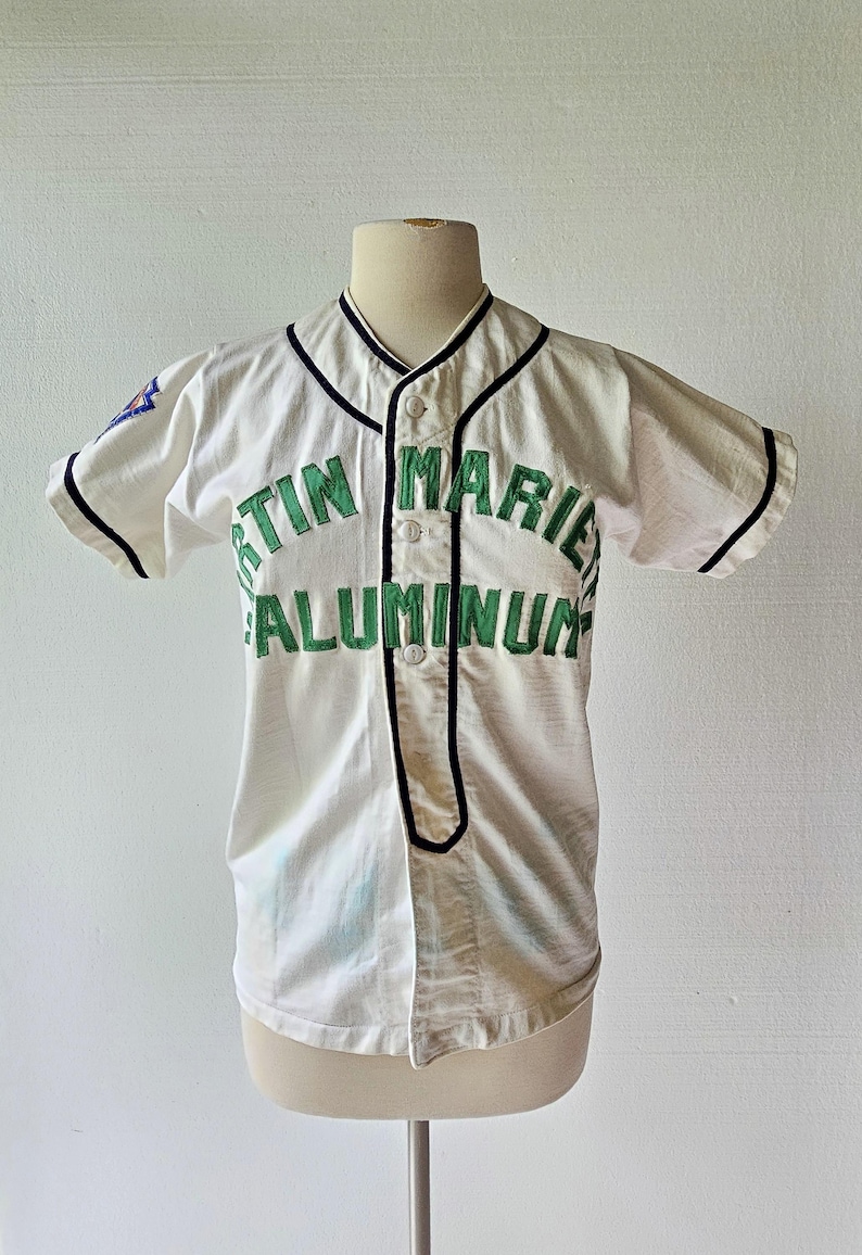 May include: White baseball jersey with black trim and green lettering that reads "TIN MARIE ALUMINUM". The jersey has short sleeves and a button-up front. A small patch is visible on the left sleeve.