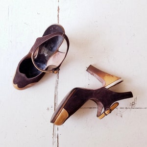 May include: A pair of brown suede heeled shoes with a gold-colored heel. One shoe has a strap across the ankle and the other has a buckle strap.