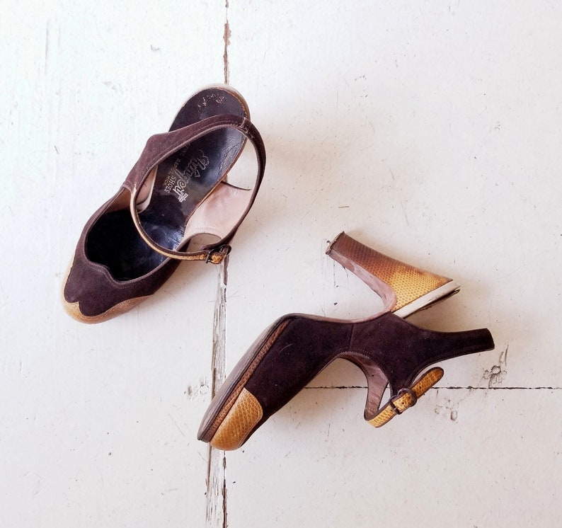 May include: A pair of brown suede heeled shoes with a gold-colored heel. One shoe has a strap across the ankle and the other has a buckle strap.