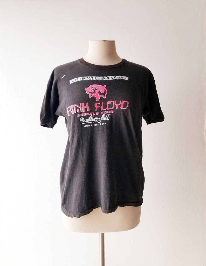 K&ouml;nnte beinhalten: Schwarzes T-Shirt mit einem pinkfarbenen Grafikdruck eines Schweins und dem Text "Pink Floyd Animals Tour" mit dem Datum "June 18 1977". Der Text "Super Bowl of Rock K-Rock 2" ist &uuml;ber dem Grafikdruck gedruckt.