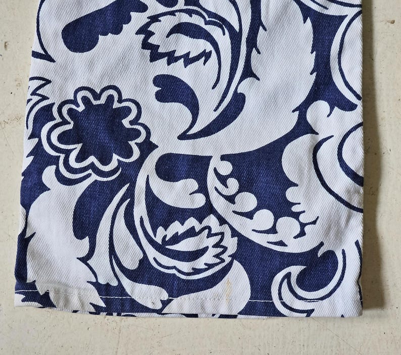 May include: Close-up of white fabric with a navy blue floral pattern. The design features stylized leaves and flower shapes. The fabric has a visible texture and a clean hemline.