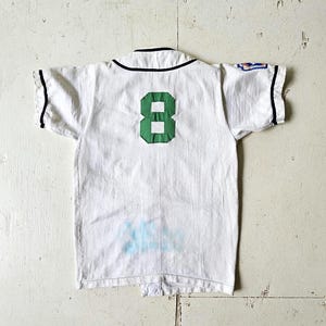 May include: White baseball jersey with a green number 8 on the back. The jersey has black trim on the collar and sleeves. There is a small patch on the right sleeve. The jersey appears to be vintage.