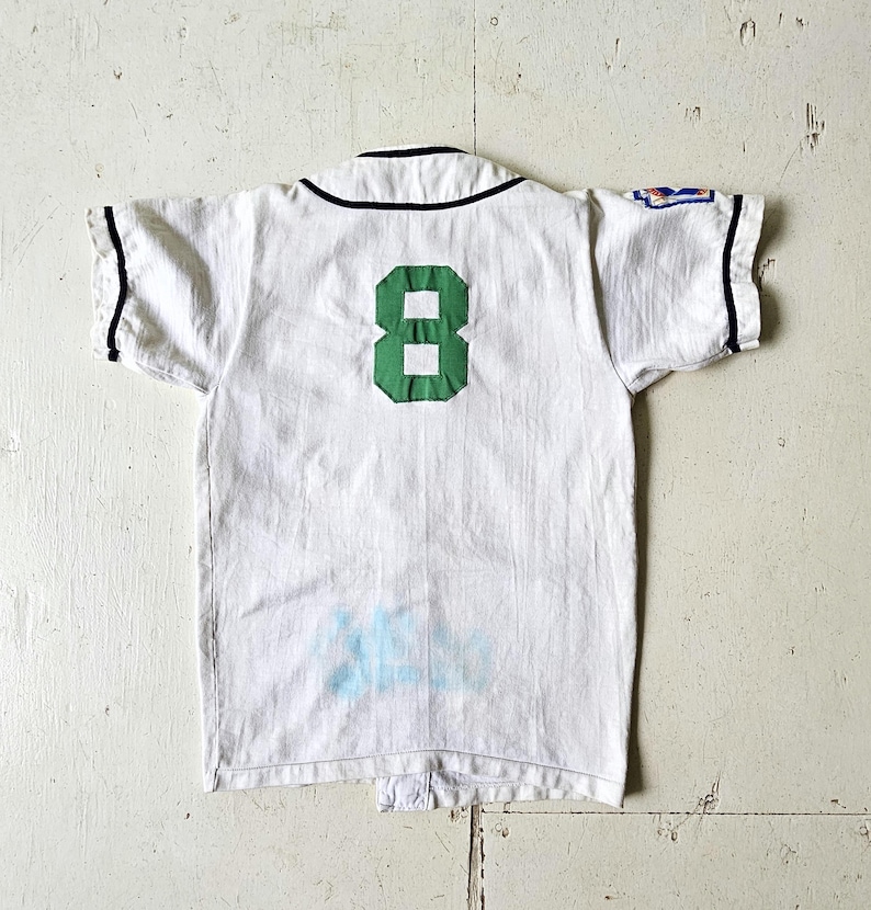 May include: White baseball jersey with a green number 8 on the back. The jersey has black trim on the collar and sleeves. There is a small patch on the right sleeve. The jersey appears to be vintage.