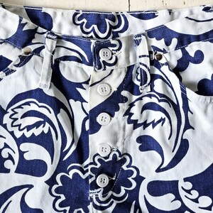 May include: Close-up of the waistband and button closure of white denim shorts with a navy blue floral pattern. The shorts have white buttons and a classic five-pocket design. The fabric is a medium weight.