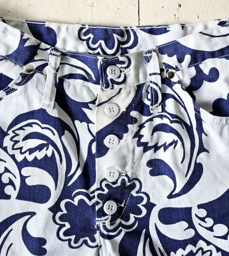 May include: Close-up of the waistband and button closure of white denim shorts with a navy blue floral pattern. The shorts have white buttons and a classic five-pocket design. The fabric is a medium weight.