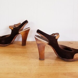 May include: A pair of brown suede heeled shoes with a strap and a wooden heel. The shoes have a contrasting light brown patch on the toe.