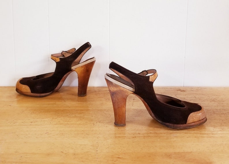 May include: A pair of brown suede heeled shoes with a strap and a wooden heel. The shoes have a contrasting light brown patch on the toe.