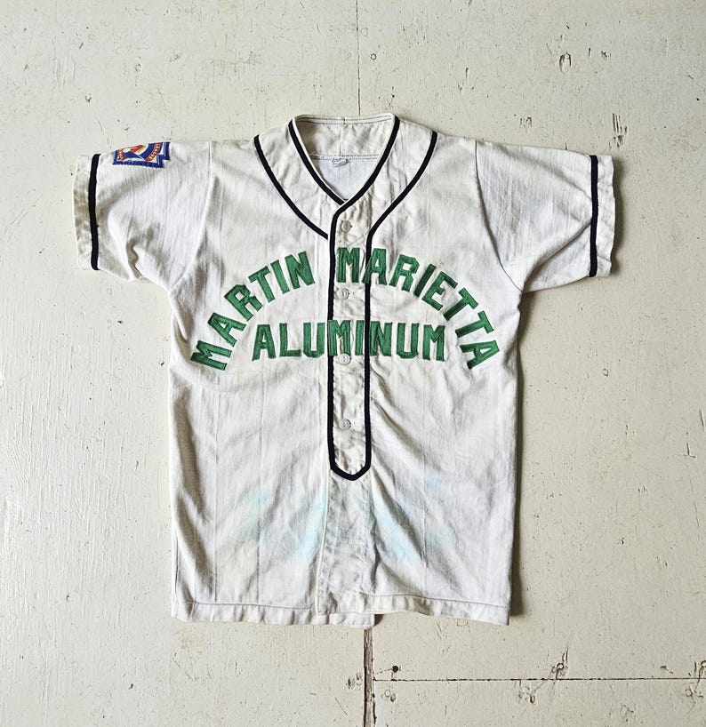 A vintage 1950s white, short sleeve, button-up baseball jersey with navy blue trim and MARTIN MARIETTA ALUMINUM in green lettering on the front; Little League patch on right sleeve.