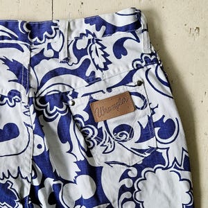 May include: Close-up of white denim pants with a blue floral pattern. The pants have a brown leather Wrangler patch on the back pocket. The design is a repeating pattern of stylized flowers and leaves.