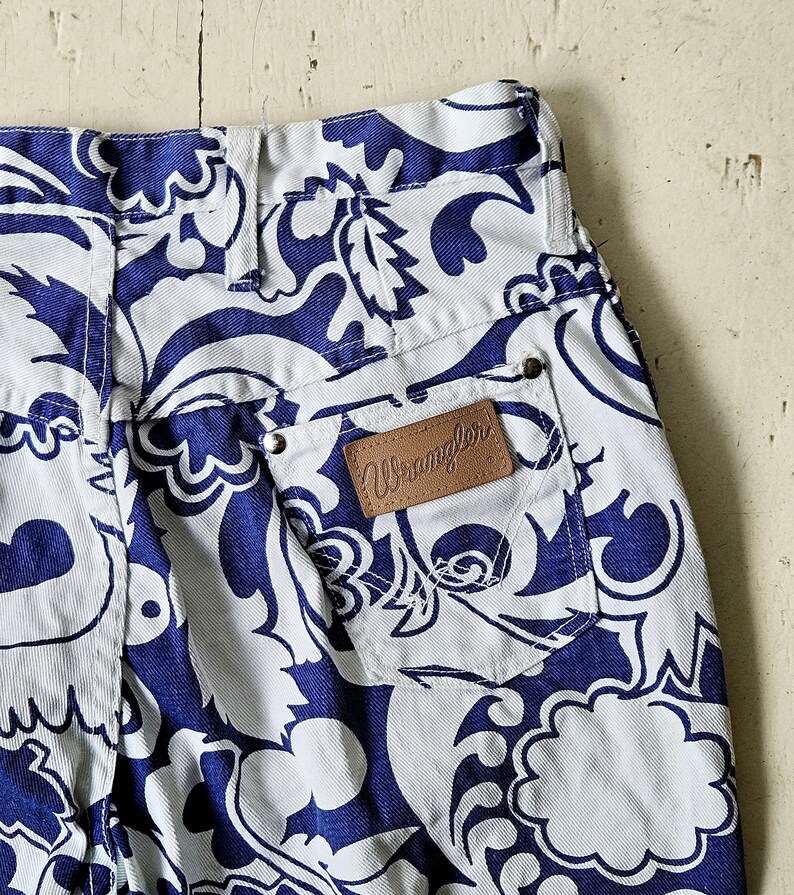 May include: Close-up of white denim pants with a blue floral pattern. The pants have a brown leather Wrangler patch on the back pocket. The design is a repeating pattern of stylized flowers and leaves.