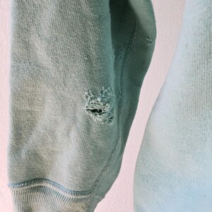 Op de afbeelding: Close-up van een versleten, lichtblauwe sweatshirtmouw. De mouw heeft een geribbelde manchet en een gat bij de elleboog. De stof vertoont tekenen van slijtage en vervaging, met een zachte, getextureerde uitstraling.