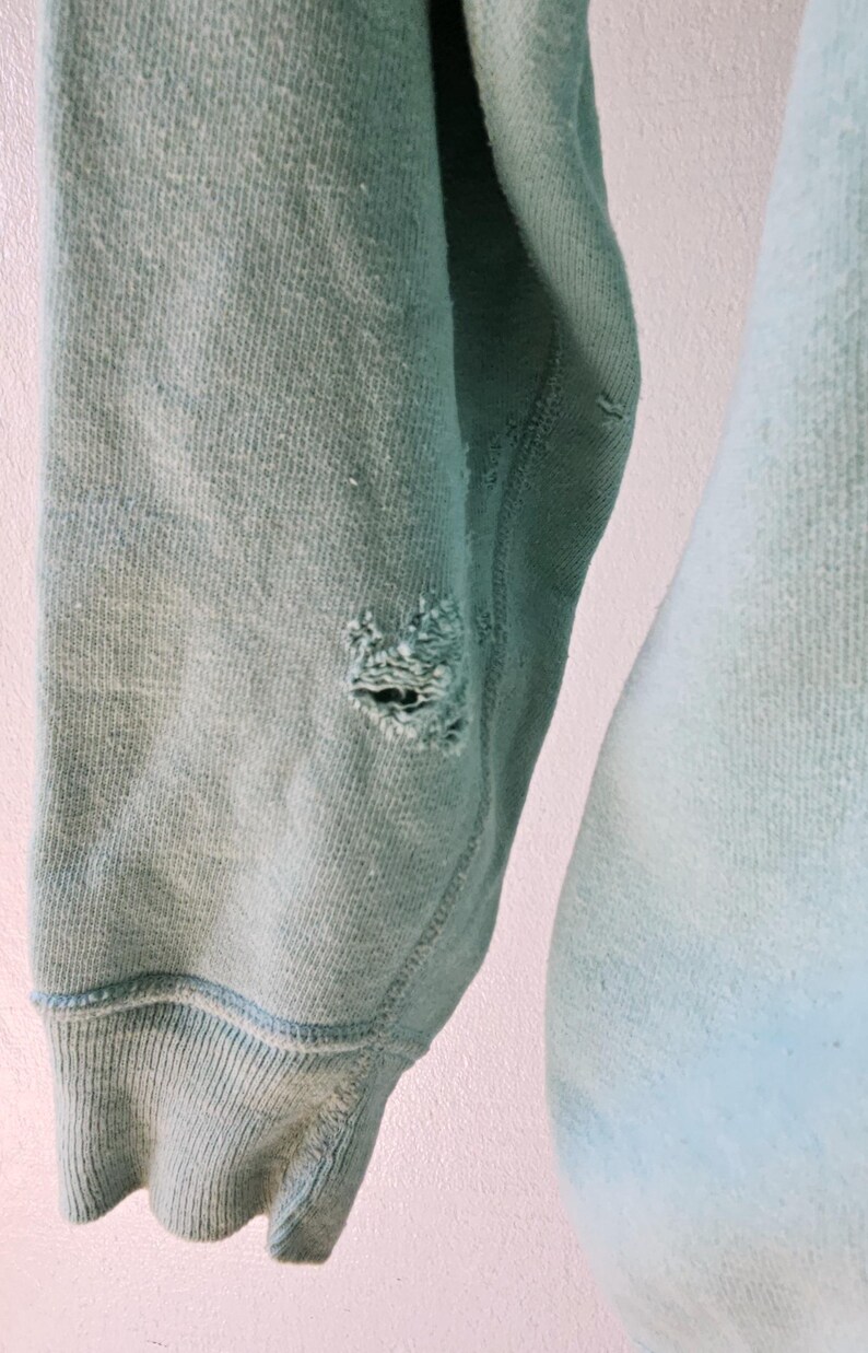 Op de afbeelding: Close-up van een versleten, lichtblauwe sweatshirtmouw. De mouw heeft een geribbelde manchet en een gat bij de elleboog. De stof vertoont tekenen van slijtage en vervaging, met een zachte, getextureerde uitstraling.