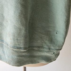 Op de afbeelding: Een vervaagde, lichtgroene sweatshirt met een geribbelde zoom en manchetten. De stof vertoont tekenen van slijtage, waaronder kleine gaatjes en verkleuring. De sweatshirt wordt op een mannequin gepresenteerd.