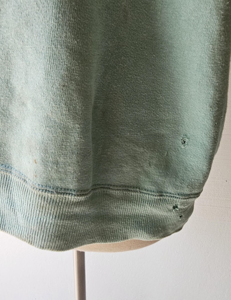 Op de afbeelding: Een vervaagde, lichtgroene sweatshirt met een geribbelde zoom en manchetten. De stof vertoont tekenen van slijtage, waaronder kleine gaatjes en verkleuring. De sweatshirt wordt op een mannequin gepresenteerd.