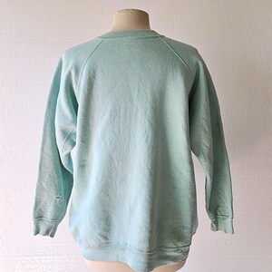 Op de afbeelding: Een vintage, lichtblauw sweatshirt met een ronde hals en lange mouwen. Het sweatshirt vertoont tekenen van slijtage, waaronder kleine gaatjes bij de manchetten. Het kledingstuk wordt op een mannequin getoond.