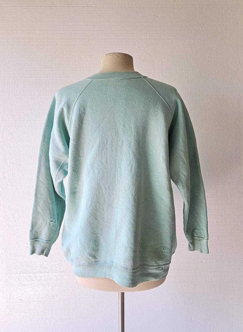 Op de afbeelding: Een vintage, lichtblauw sweatshirt met een ronde hals en lange mouwen. Het sweatshirt vertoont tekenen van slijtage, waaronder kleine gaatjes bij de manchetten. Het kledingstuk wordt op een mannequin getoond.