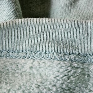 Op de afbeelding: Close-up van de halslijn van een lichtblauw sweatshirt. De geribbelde kraag, met verticale lijnen, is vastgenaaid aan de zachtere fleece stof eronder. De afbeelding benadrukt de textuur en constructie van het kledingstuk.