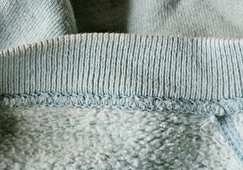 Op de afbeelding: Close-up van de halslijn van een lichtblauw sweatshirt. De geribbelde kraag, met verticale lijnen, is vastgenaaid aan de zachtere fleece stof eronder. De afbeelding benadrukt de textuur en constructie van het kledingstuk.