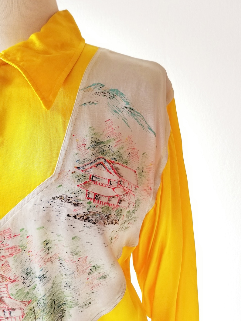 Puede incluir: Una camisa amarilla de manga larga con un panel blanco que presenta un dise&ntilde;o de paisaje de inspiraci&oacute;n japonesa. El dise&ntilde;o incluye una cordillera, una casa y &aacute;rboles en tonos de verde, rojo y azul.