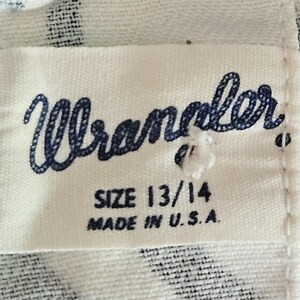 May include: Close-up of a white fabric tag with the word "Wrangler" in navy blue script. Below the brand name are the size "13/14" and the text "MADE IN U.S.A." The tag is sewn onto a patterned fabric.