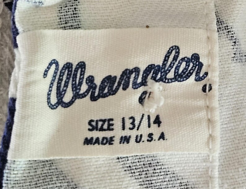May include: Close-up of a white fabric tag with the word "Wrangler" in navy blue script. Below the brand name are the size "13/14" and the text "MADE IN U.S.A." The tag is sewn onto a patterned fabric.