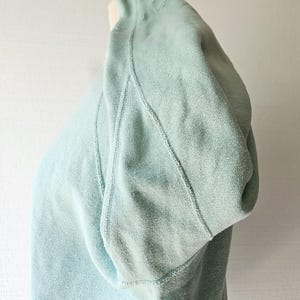 Op de afbeelding: Een lichtblauw sweatshirt met een capuchon. Het sweatshirt is gemaakt van een zachte, getextureerde stof. De naden zijn zichtbaar en de capuchon is omgeslagen. De achtergrond is een effen witte muur.