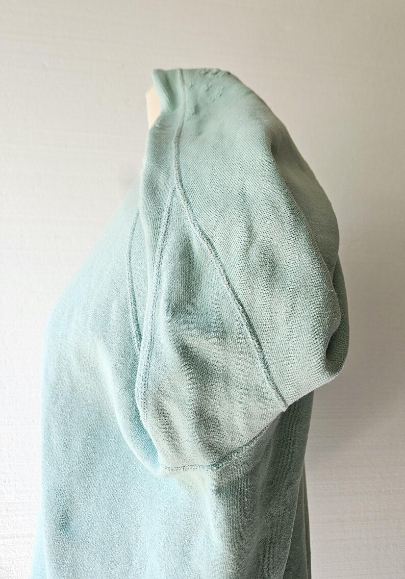 Op de afbeelding: Een lichtblauw sweatshirt met een capuchon. Het sweatshirt is gemaakt van een zachte, getextureerde stof. De naden zijn zichtbaar en de capuchon is omgeslagen. De achtergrond is een effen witte muur.