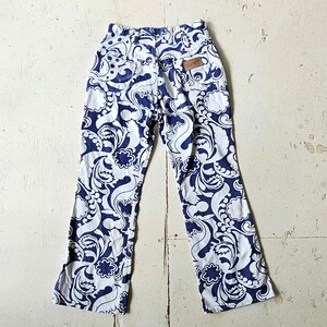 May include: White and navy blue patterned pants with a high waist and flared legs. The pants feature a repeating floral and swirl design. A small brown leather tag with the word "Wrangler" is visible on the back.