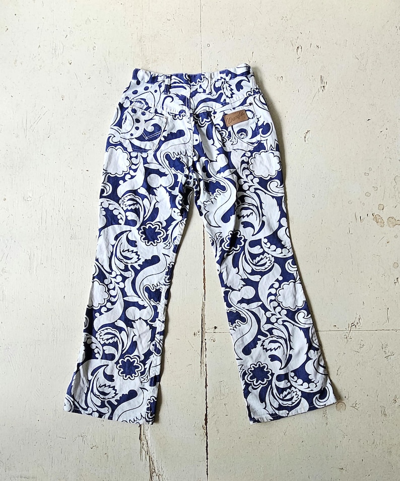 May include: White and navy blue patterned pants with a high waist and flared legs. The pants feature a repeating floral and swirl design. A small brown leather tag with the word "Wrangler" is visible on the back.