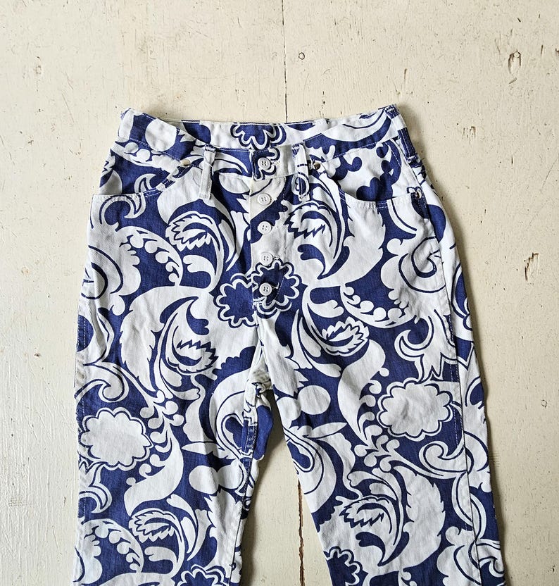 May include: High-waisted pants with a white and navy blue floral pattern. The pants have a button fly and two front pockets. The fabric has a swirling, paisley-like design. The pants are made of a lightweight material.