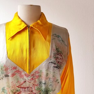 Puede incluir: Una chaqueta de sat&eacute;n amarilla con cierre de cremallera y un panel blanco con un dise&ntilde;o pintado a mano de una pagoda japonesa y un paisaje.