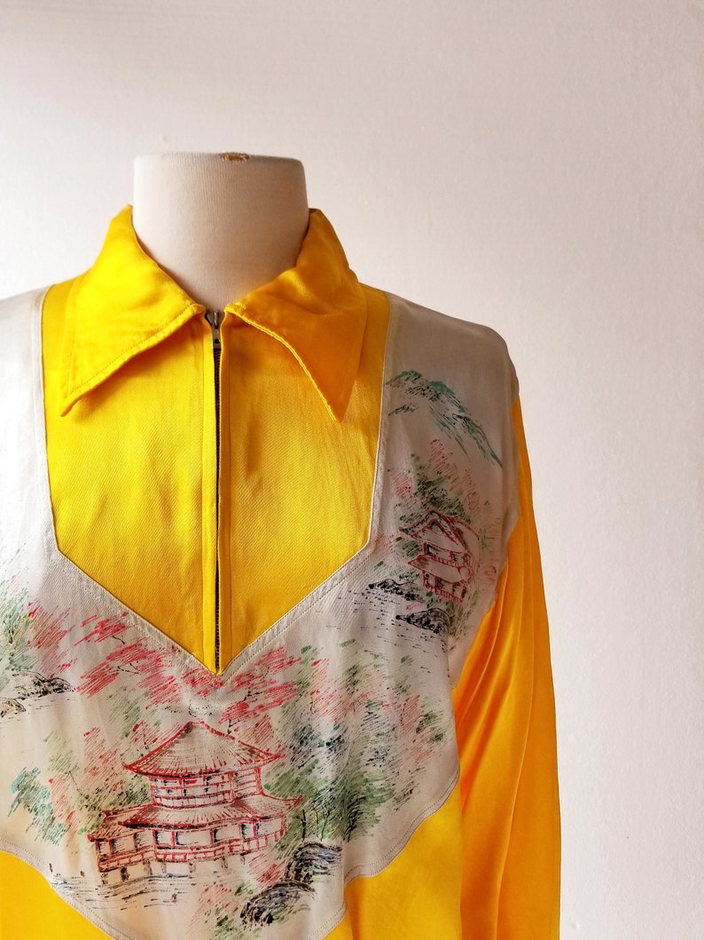 Puede incluir: Una chaqueta de sat&eacute;n amarilla con cierre de cremallera y un panel blanco con un dise&ntilde;o pintado a mano de una pagoda japonesa y un paisaje.
