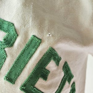May include: Close-up of a cream-colored fabric with green embroidered letters. The letters are stitched with a textured, ribbed design. The fabric has a slight texture and a small imperfection.