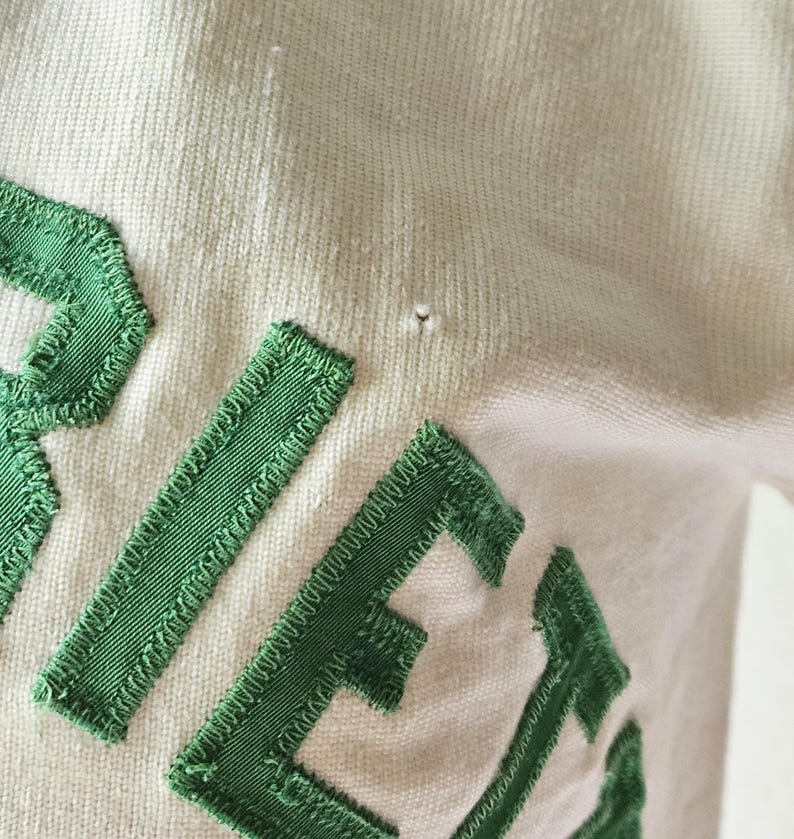 May include: Close-up of a cream-colored fabric with green embroidered letters. The letters are stitched with a textured, ribbed design. The fabric has a slight texture and a small imperfection.