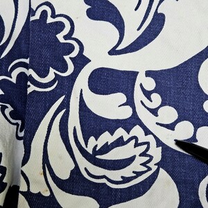May include: Close-up of a fabric with a repeating pattern of white floral designs on a navy blue background. The fabric has a textured appearance, with visible weave details. The design includes stylized flowers and swirling shapes.
