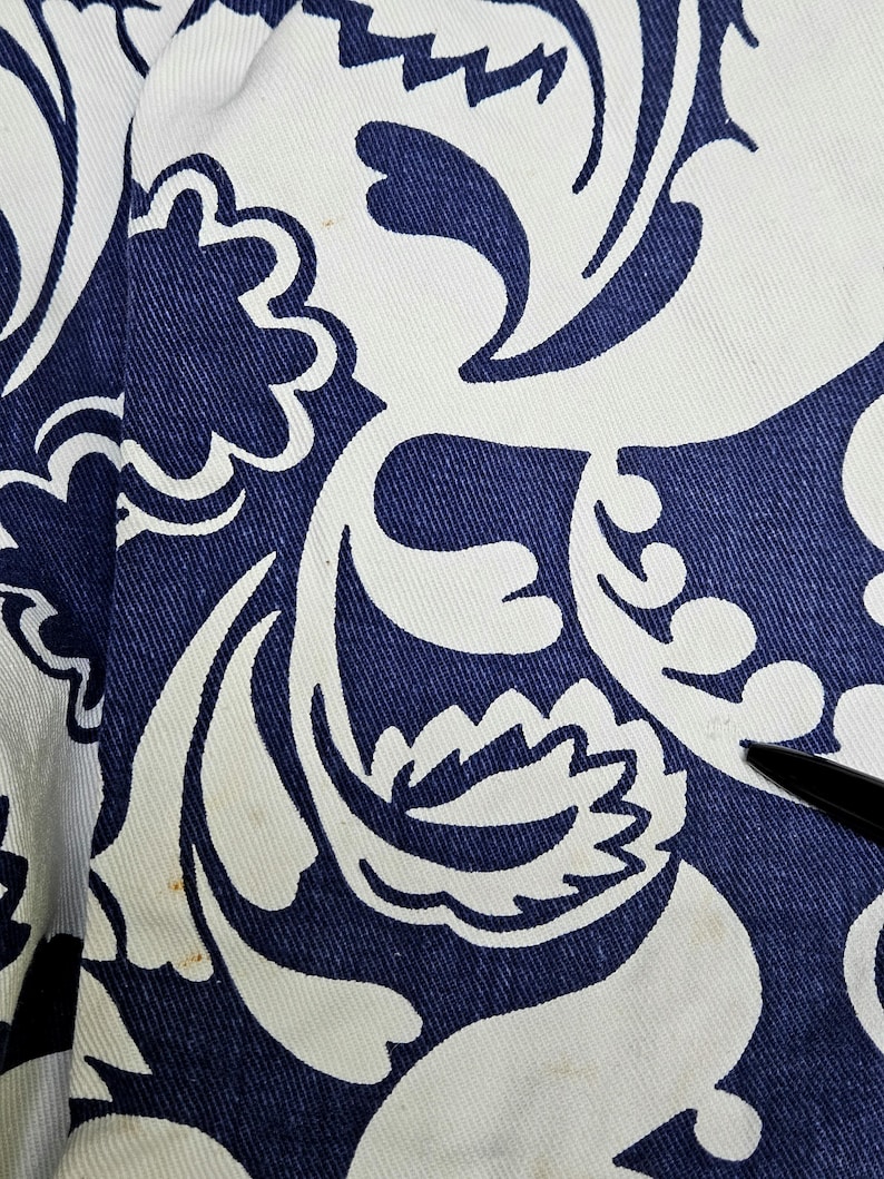 May include: Close-up of a fabric with a repeating pattern of white floral designs on a navy blue background. The fabric has a textured appearance, with visible weave details. The design includes stylized flowers and swirling shapes.
